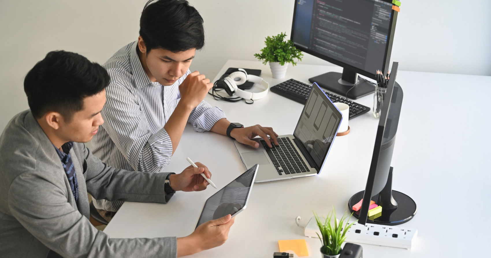 Two individuals collaborating at a desk using a laptop and a tablet with a stylus, while a monitor displaying code and office items like headphones and a plant are visible in the background.