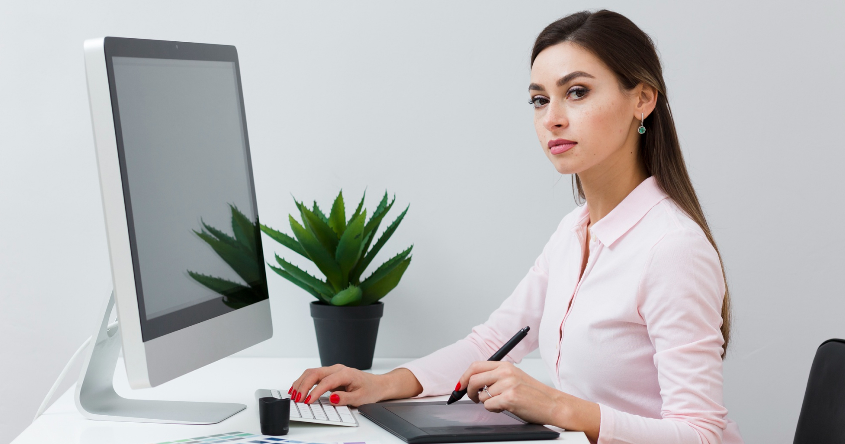 Individual seated at a desk using a stylus on a graphic tablet next to a desktop computer, with a small potted plant beside the monitor in a clean workspace.
