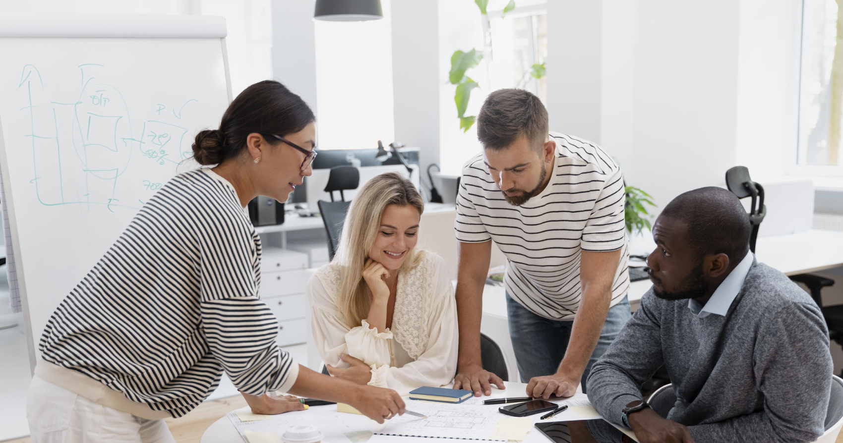 Four individuals gathered around a table collaborating on documents, with one pointing to a plan on paper while others review notes and a smartphone in a bright office setting.