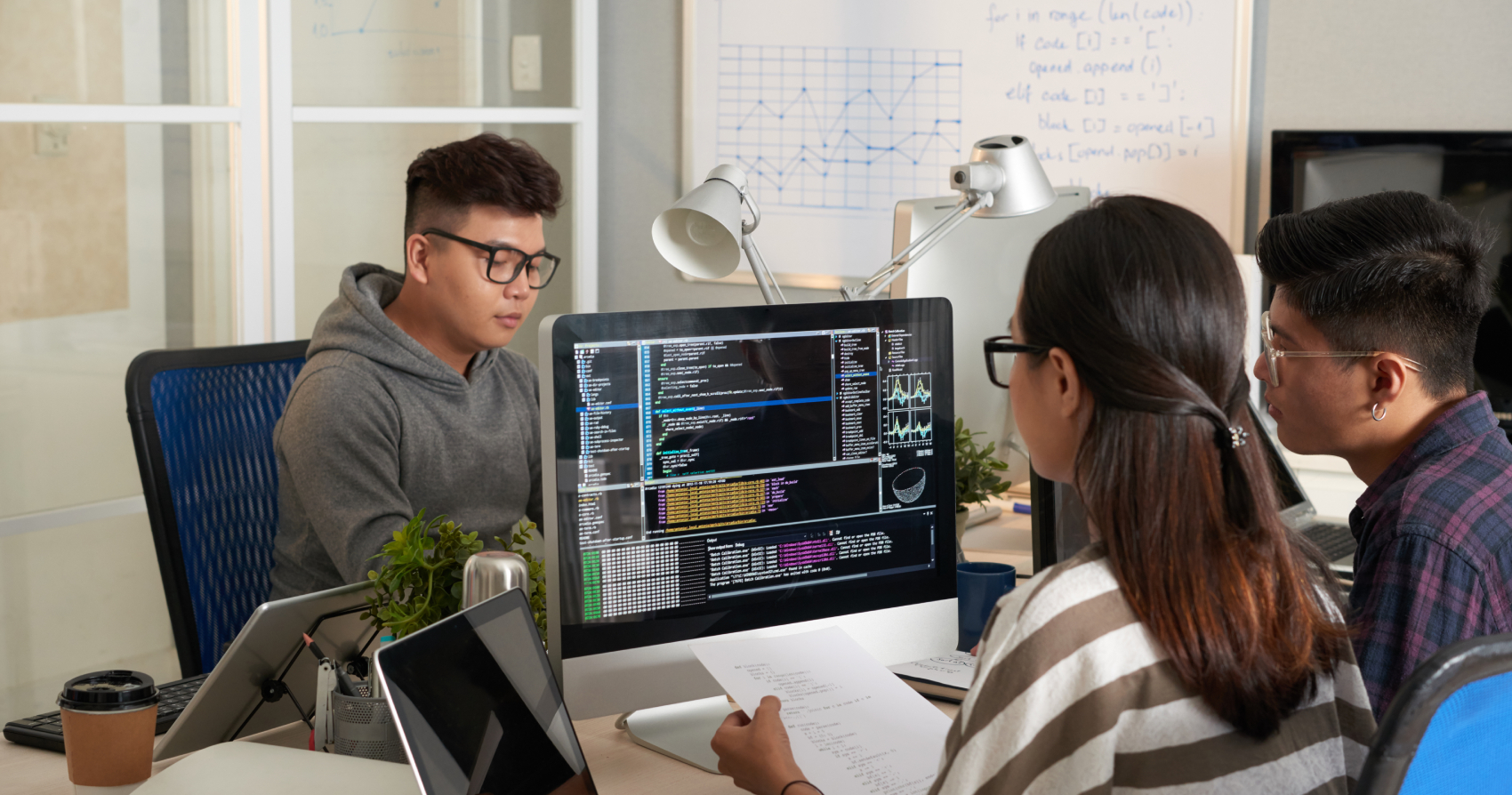 Three individuals collaborating around a desktop computer displaying code, with one reviewing printed notes and others observing the screen in a modern office workspace.