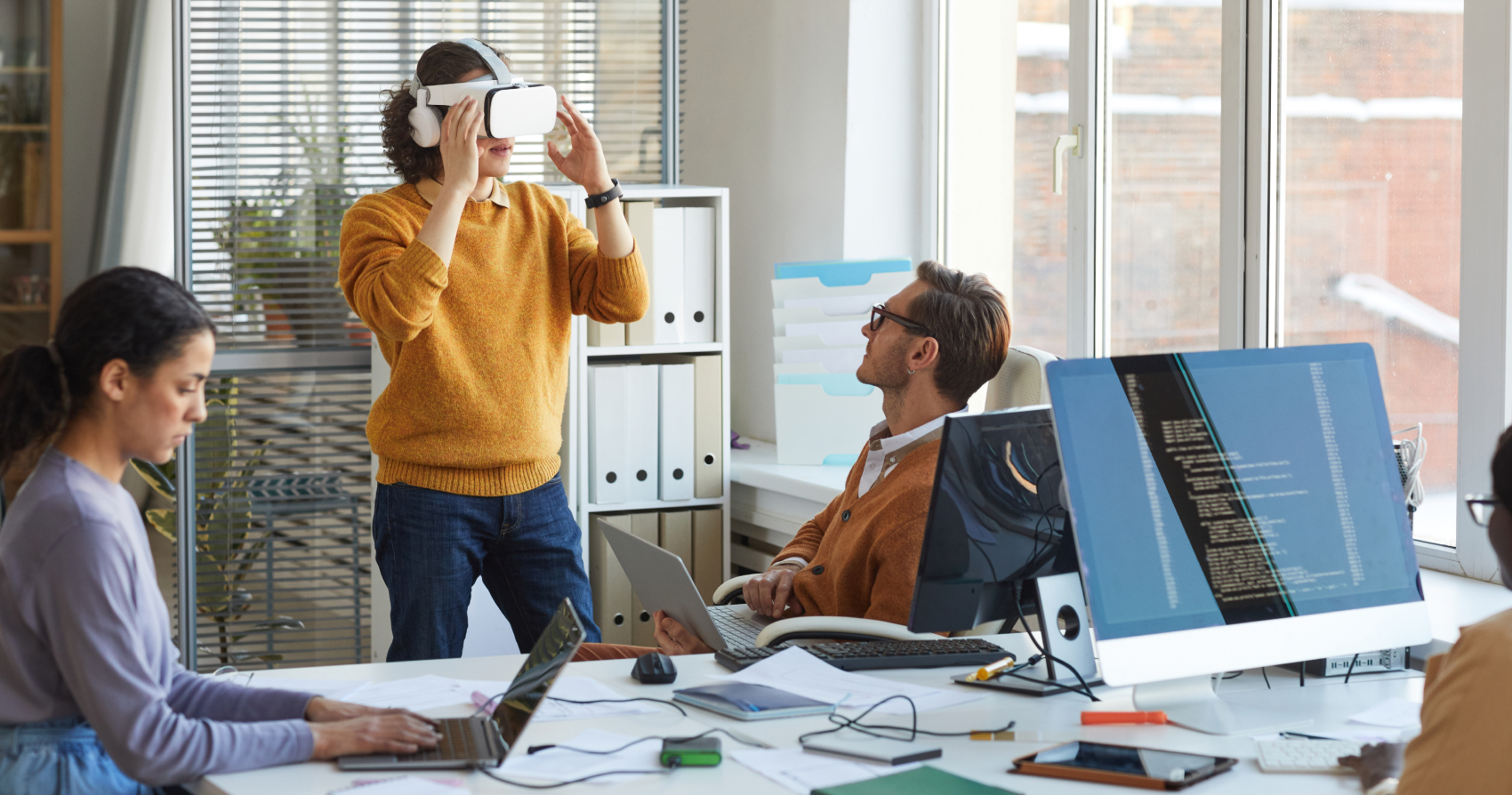 Office environment where a person is standing and using a virtual reality headset while others work on laptops and desktop computers, with code displayed on a monitor.