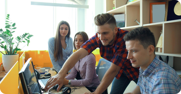 Group of four professionals gathered around a desk, collaborating while looking at a laptop, with one person pointing at the screen and others observing in a modern office environment.