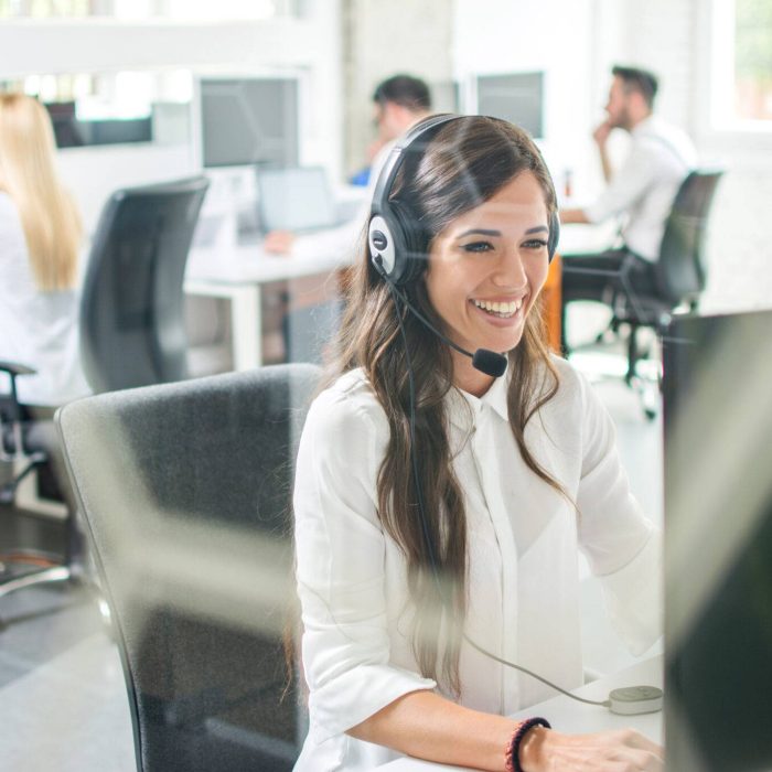 Customer support agent wearing a headset and working at a computer in a bright modern office with colleagues in the background.